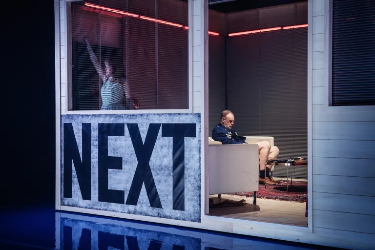 Flicka vid fönster till vänster. Man i soffa till höger. De sitter inuti ett hus på scenen.