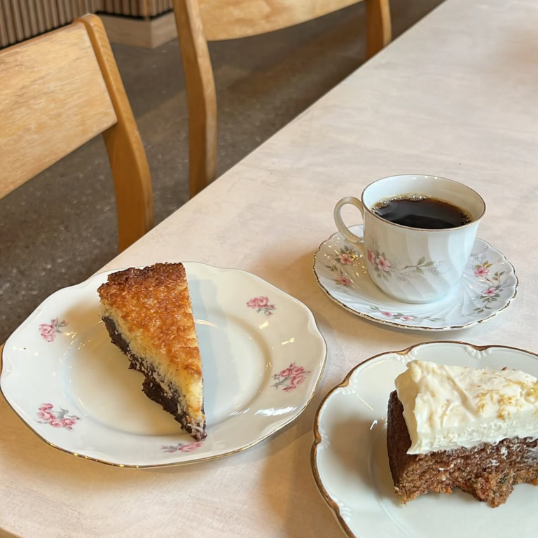 Två vackert dekorerade tallrikar med kakor – en med chokladkaka med vit glasyr och en med mörk paj med ett gyllene kokostäcke. En kopp svart kaffe i en blommig porslinskopp står bredvid på ett ljust dukat bord. Två trästolar syns i bakgrunden.
