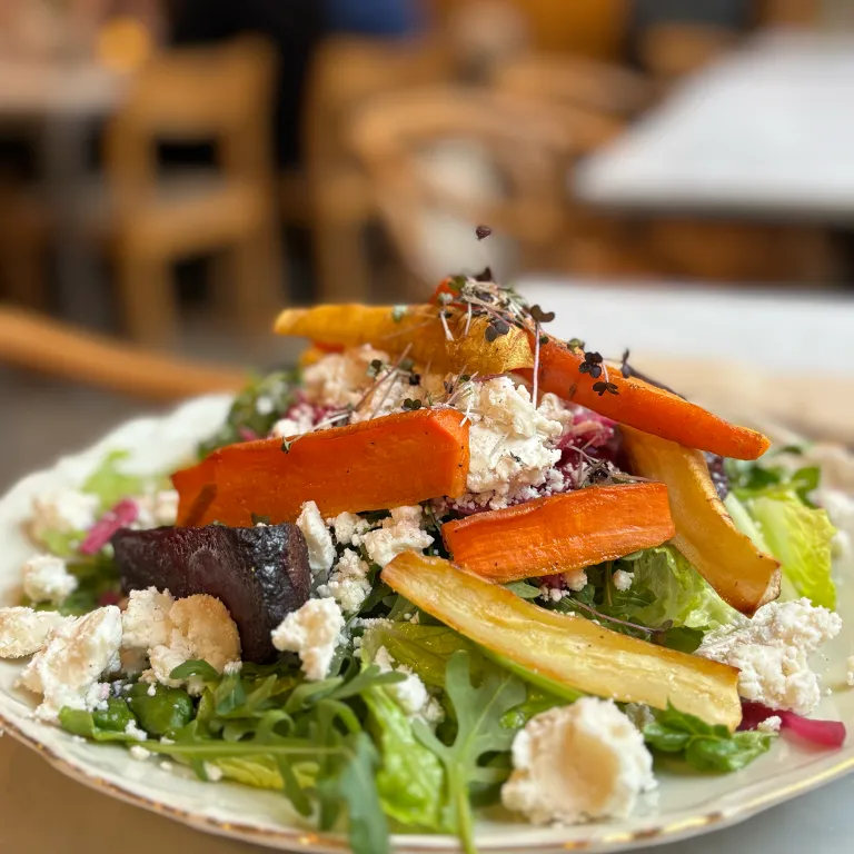 En färgglad sallad toppad med rostade rotfrukter, såsom morötter och rödbetor, samt smulad fetaost och späda skott. Rätten serveras på en rustik tallrik och står på ett bord i en inbjudande caféatmosfär med trästolar i bakgrunden.