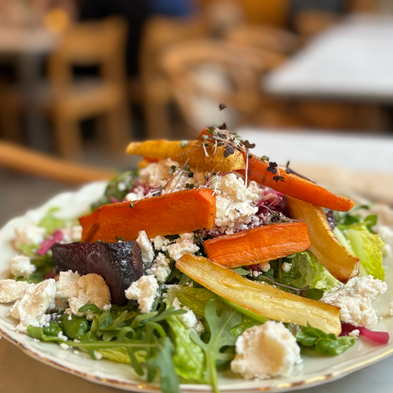 En färgglad sallad toppad med rostade rotfrukter, såsom morötter och rödbetor, samt smulad fetaost och späda skott. Rätten serveras på en rustik tallrik och står på ett bord i en inbjudande caféatmosfär med trästolar i bakgrunden.