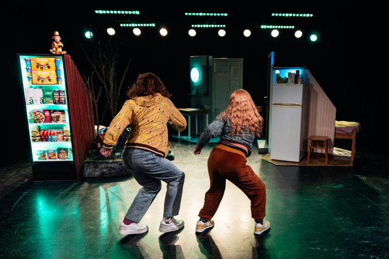 Två personer med ryggen mot kameran dansar eller rör sig energiskt på en teaterscen. Till vänster syns en upplyst hylla med matvaror som i en mataffär, och till höger ett kylskåp, en säng och andra rekvisitadetaljer som ger scenen en hemmiljö.