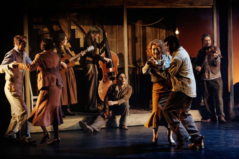 En dynamisk scen fylld av musik och rörelse – två par dansar intensivt medan musiker spelar i bakgrunden. En kvinna och en man dansar passionerat i förgrunden, och scenen genomsyras av energi och dramatik.