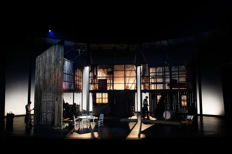 En stämningsfull scenbild av en teateruppsättning, där ett tvåvåningshus med rustika träväggar och gamla fönster dominerar scenen. Skådespelare rör sig genom det dunkla ljuset och skuggor spelar över golvet.