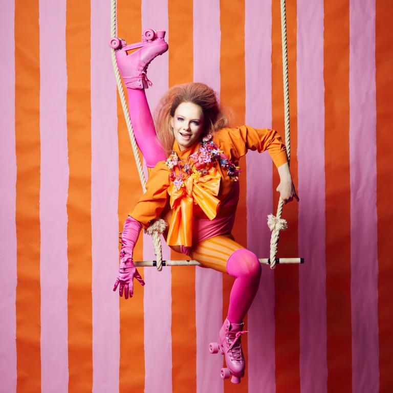 Klara G. En artist i färgglad cirkuskostym poserar akrobatiskt på en trapets framför en bakgrund med vertikala rosa och orange ränder. Hen bär rosa rullskridskor, rosa strumpbyxor, orange tröja med en stor orange rosett och blommor på bröstet, samt rosa handskar. En bensträckning upp i luften visar på flexibilitet och glädje i uttrycket.