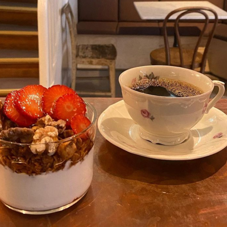 På en kopparbänk står en antik kaffekopp med små rosa blommor med svart kaffe i. Bredvid står ett glas med yoghurt toppad med granola och jordgubbar 