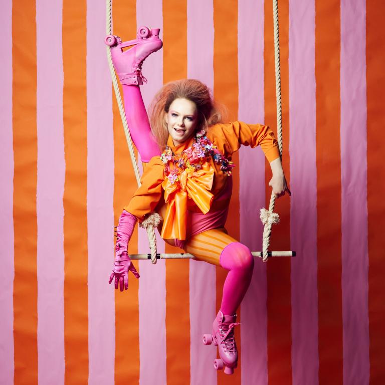 Klara G. Tjej med orange blus, rosa strumpbyxor och rullskridskor sitter på en trapets med ena benet upp i luften. Bakgrunden är organgerosa ränder