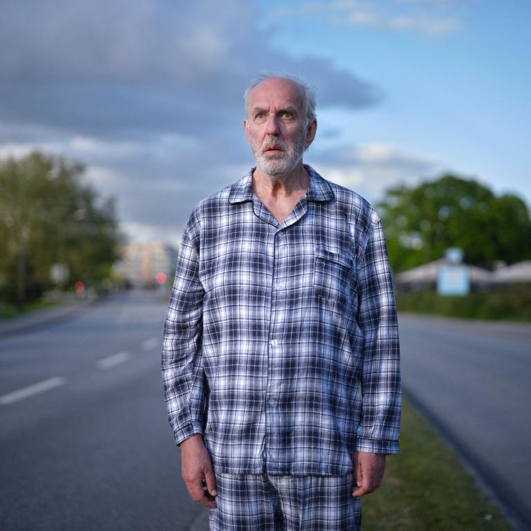 En äldre man står i pyjamas mitt i vägen och ser förbryllad ut.