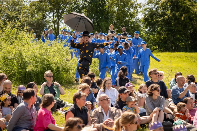  Foto utomhus i soligt väder. Vuxen skådespelare iklädd svart pyjamas, svart hatt och svart paraply dansar ner för en slänt, förbi publik. Bakom honom många barn i likadana blå pyjamas.