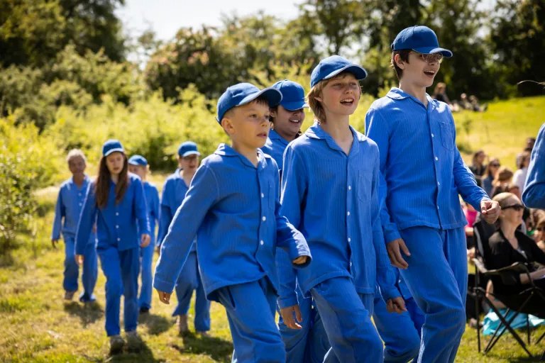  Foto utomhus i soligt väder. Barn i likadana blå pyjamas dansar och sjunger förbi publik som sitter i gräset.