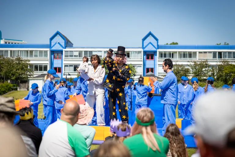  Foto utomhus i soligt väder. Många barn i likadana blå pyjamas på scen tillsammans med två vuxna skådespelare. Den ena i vit pyjamas, den andra i svart med gula stjärnor. De har varsin liten docka av sig själva i händerna. I bakgrunden många barn i blå pyjamas, i förgrunden publik som sitter i gräset