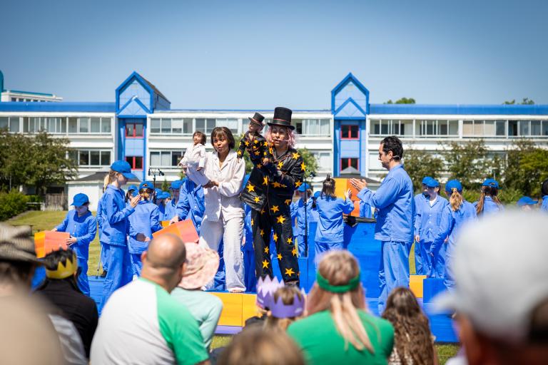  Foto utomhus i soligt väder. Många barn i likadana blå pyjamas på scen tillsammans med två vuxna skådespelare. Den ena i vit pyjamas, den andra i svart med gula stjärnor. De har varsin liten docka av sig själva i händerna. I bakgrunden många barn i blå pyjamas, i förgrunden publik som sitter i gräset