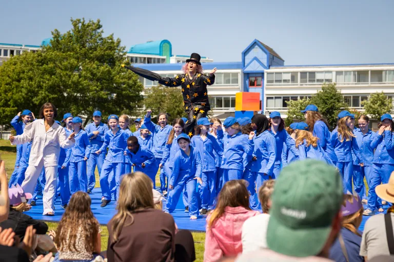 Foto från utomhusteater. Publik som sitter i gräset och en scen på marken med skådespelare och andra medverkande på. En vuxen skådespelare i svart pyjamas står på låda och sträcker ut sina händer, och ser ut att sjunga.