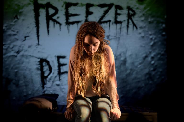 Kvinna sitter på en säng med huvudet nedböjt, bakom henne syns texten "Freezer of Death".