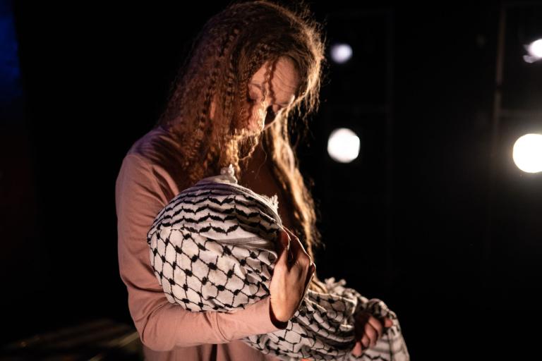 Kvinna tittar ner mot ett bylte i famnen, svept i en keffiyeh, med starka lampor i bakgrunden.
