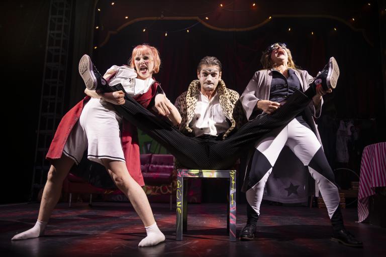 Sara P. Borgström Tre artister gör humoristiskt nummer där en person sitter på stol med benen utsträckta.