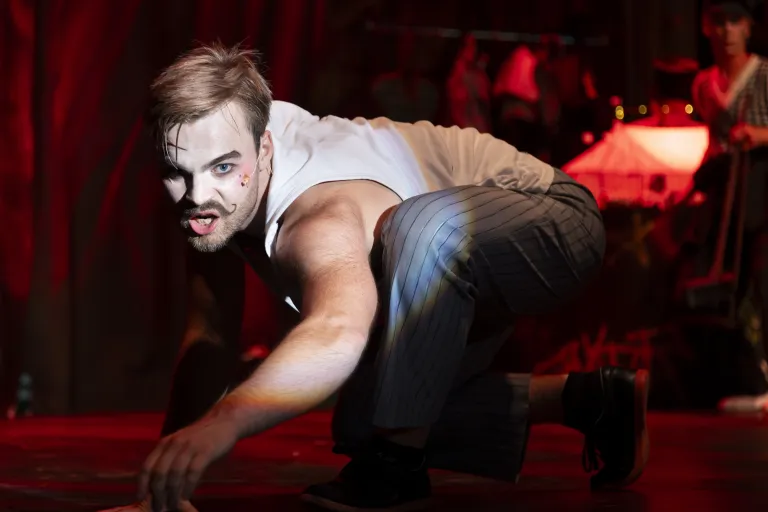 Artist med målad mustasch kryper på scenen under starkt rött ljus.