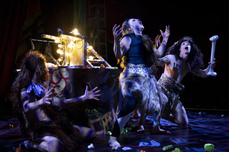 Sara P. Borgström Tre artister utklädda till stenåldersmänniskor gestikulerar dramatiskt på scen.