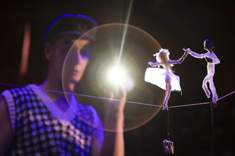 Artist håller ficklampa mot två dockor som balanserar på lina.