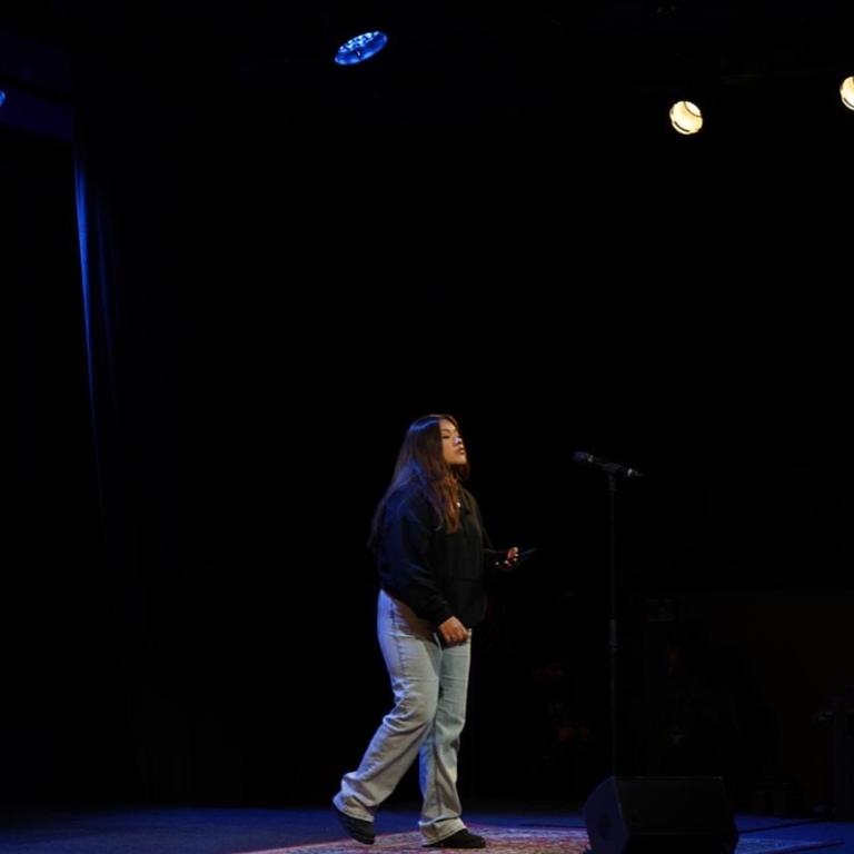 Illari Jozwikowska  Person står i jeans pch svart colleagetröja vid en miktofon på en upplyst scen.