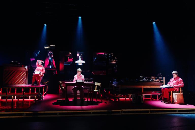 Personen sitter vid ett bord i en scenbild med rött ljus, medan två andra personer sitter på varsin upphöjd plattform.