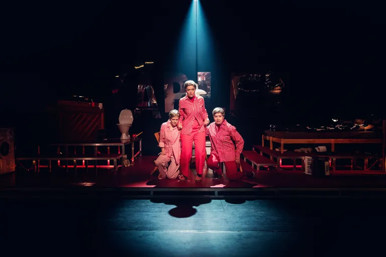Tre personer i röda och rosa kostymer står mitt på scenen under en spotlight.