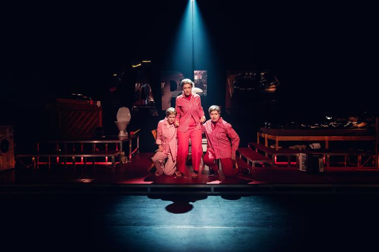 Tre personer i röda och rosa kostymer står mitt på scenen under en spotlight.