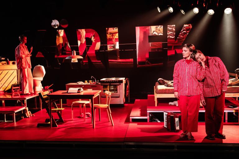 Två personer i rödrandiga kläder står nära varandra i en scenmiljö med kök och säng; en tredje person står på en upphöjd plattform.