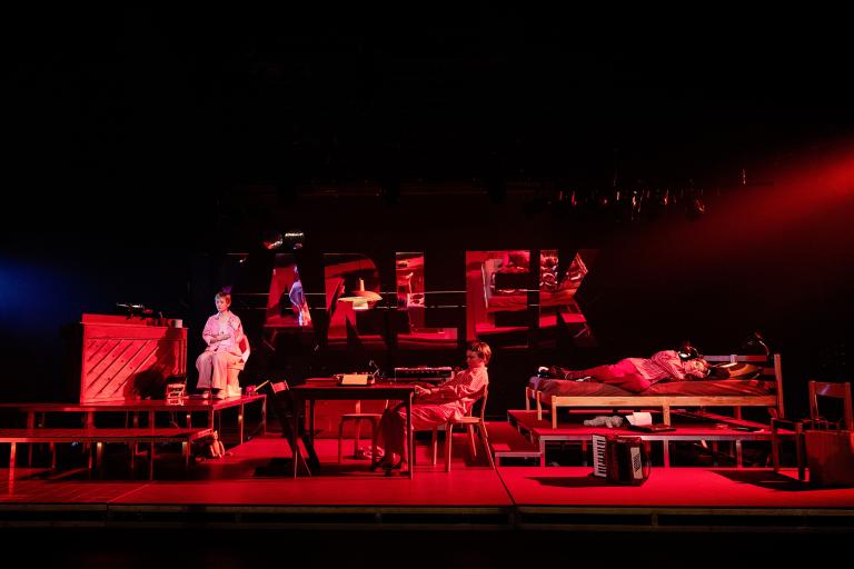 En person sitter på en upphöjd plattform och två personer befinner sig vid bord och säng i en röd scenbild.