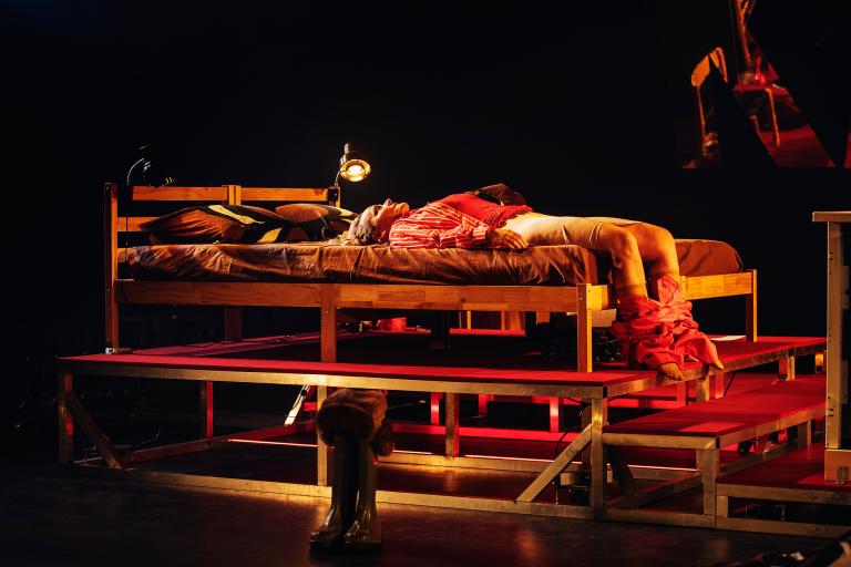 En person ligger på en säng med benen hängande ner från kanten i röd scenbelysning.