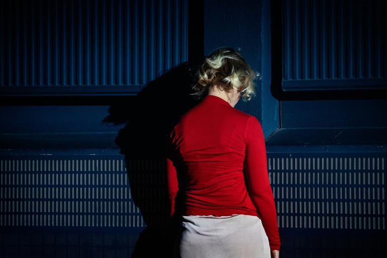 En person står vänd mot en blå vägg i en mörk scenmiljö.