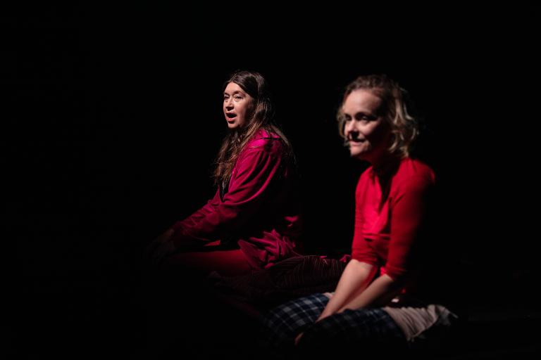 Två personer sitter i mörker, belysta av en spotlight, på en scen utan synliga möbler.