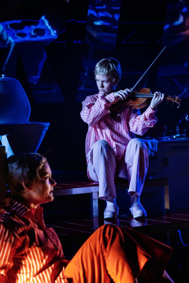 Person i rosa pyjamas sitter på plattform och spelar fiol; en annan person syns suddigt i förgrunden.