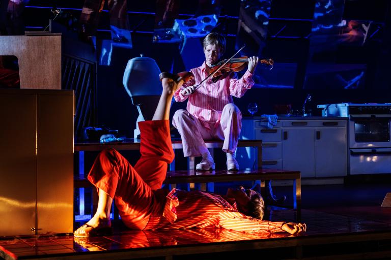 Person spelar fiol på en plattform medan en annan person ligger på golvet framför.