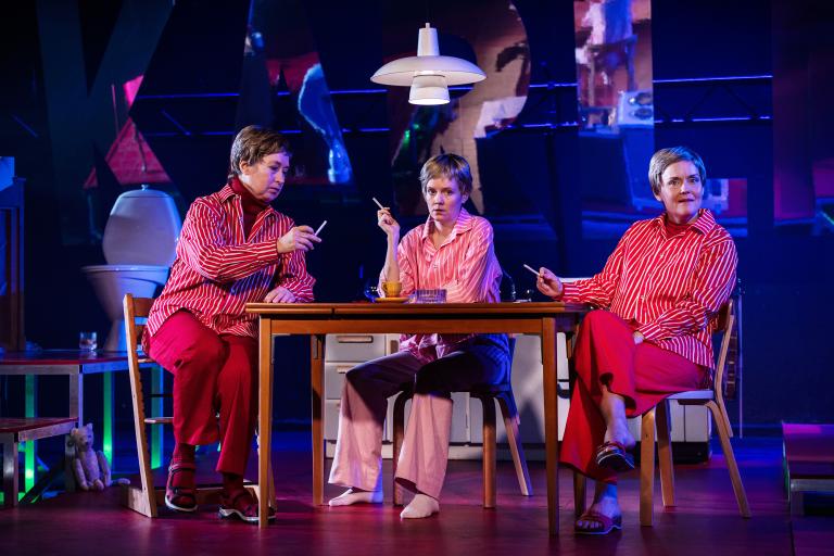 Tre personer i randiga skjortor sitter vid ett köksbord med cigaretter i händerna.