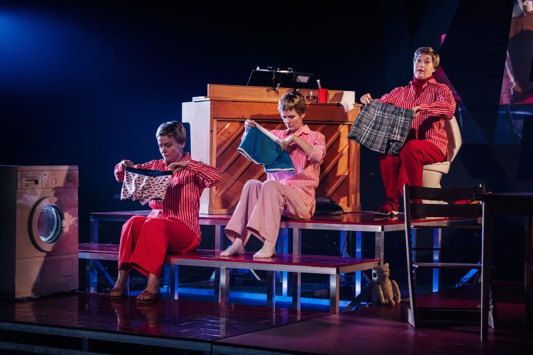 Tre personer sitter på trappsteg och håller upp olika plagg bredvid en tvättmaskin.