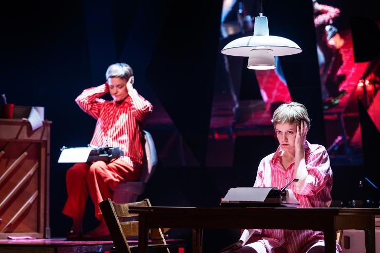 En person sitter vid ett bord med skrivmaskin medan en annan sitter bakom på en toalett.