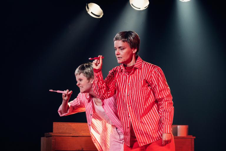 Två personer i randiga skjortor håller upp varsin rosa tandborste under stark scenbelysning.