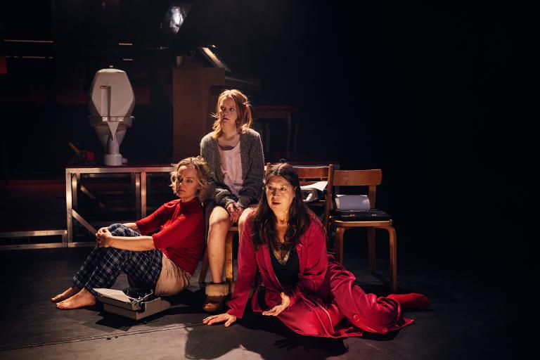 Tre personer sitter på scengolvet bredvid varandra och sjunger