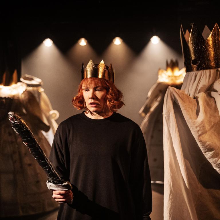 Skådespelare med krona och svarta kläder tittar på sitt svärd som hon håller i handen. Bakom syns en spökfigur med guldkrona