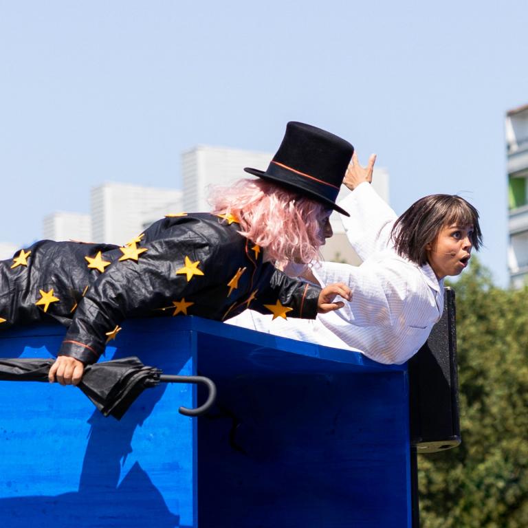 Två skådespelare ligger på en stor blå låda och låtsas flyga