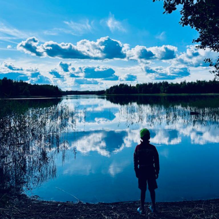 et barn står och tittar ut över en sjö. Himlen speglar sig i vattnet. träd runt om. 