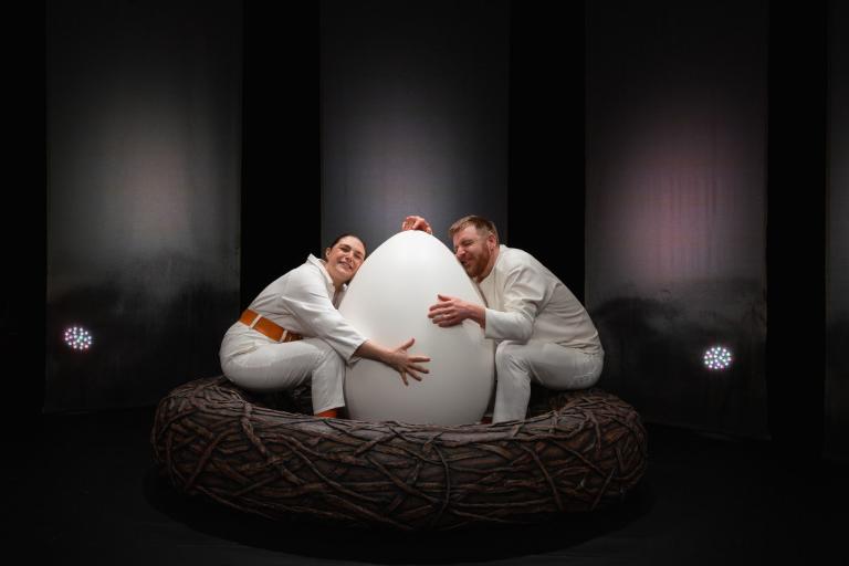Två skådespelare sitter vid ett stort vitt ägg och kramar om det. 