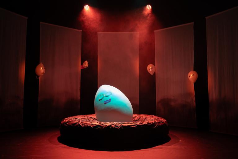 Ett stort ägg ligger i ett bo på en scen. Rött ljus och i bakgrunden syns vita ballonger med ritade arga ansikten.