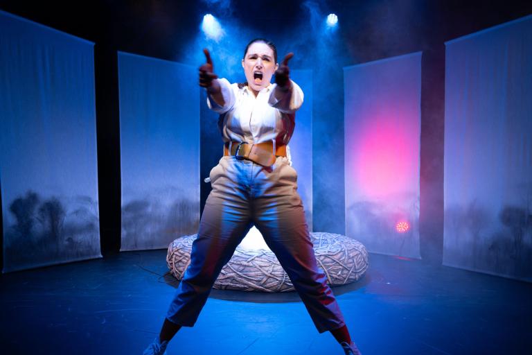 Person står bredbent i dramatiskt ljus och riktar armarna rakt fram på scenen.