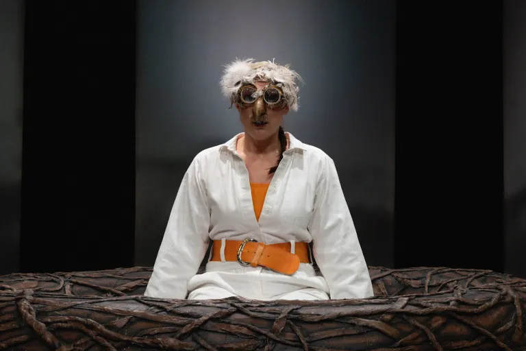 Närbild på skådespelare med fågelmask sittande i stort bo på scenen.