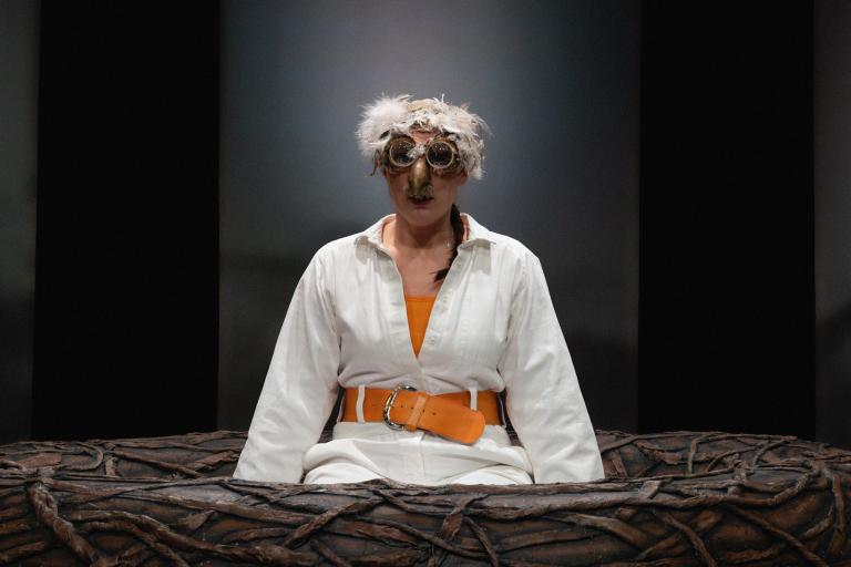 Närbild på skådespelare med fågelmask sittande i stort bo på scenen.
