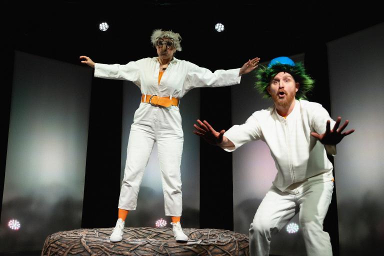 Två skådespelare i lekfulla peruker, en balanserar på boet och den andra bredvid med armarna utsträckta.