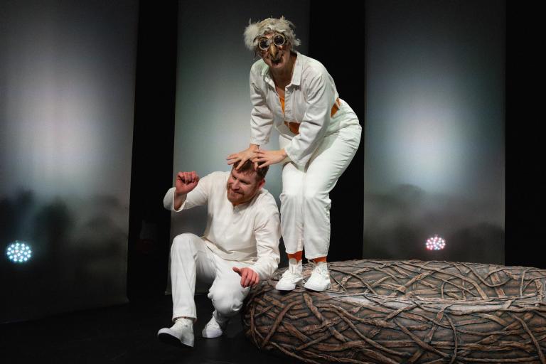 Skådespelare med fågelmask står i boet och håller händerna på en annan persons huvud.