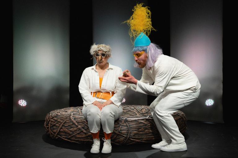 En skådespelare i turkos hatt med gul tylltoffs håller i en puderdosa. En annan i fågelmask sitter på ett stort bo.