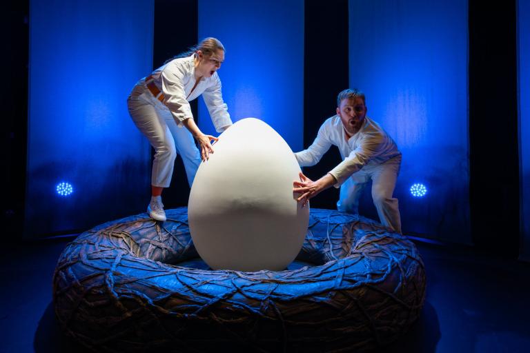 Två skådespelare står i ett stort fågelbo och håller på ett stort vitt ägg i blått scenljus.